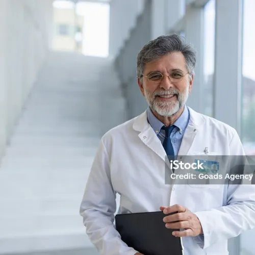 Experienced physician wearing a lab coat and holding a file, standing in a bright hospital hallway